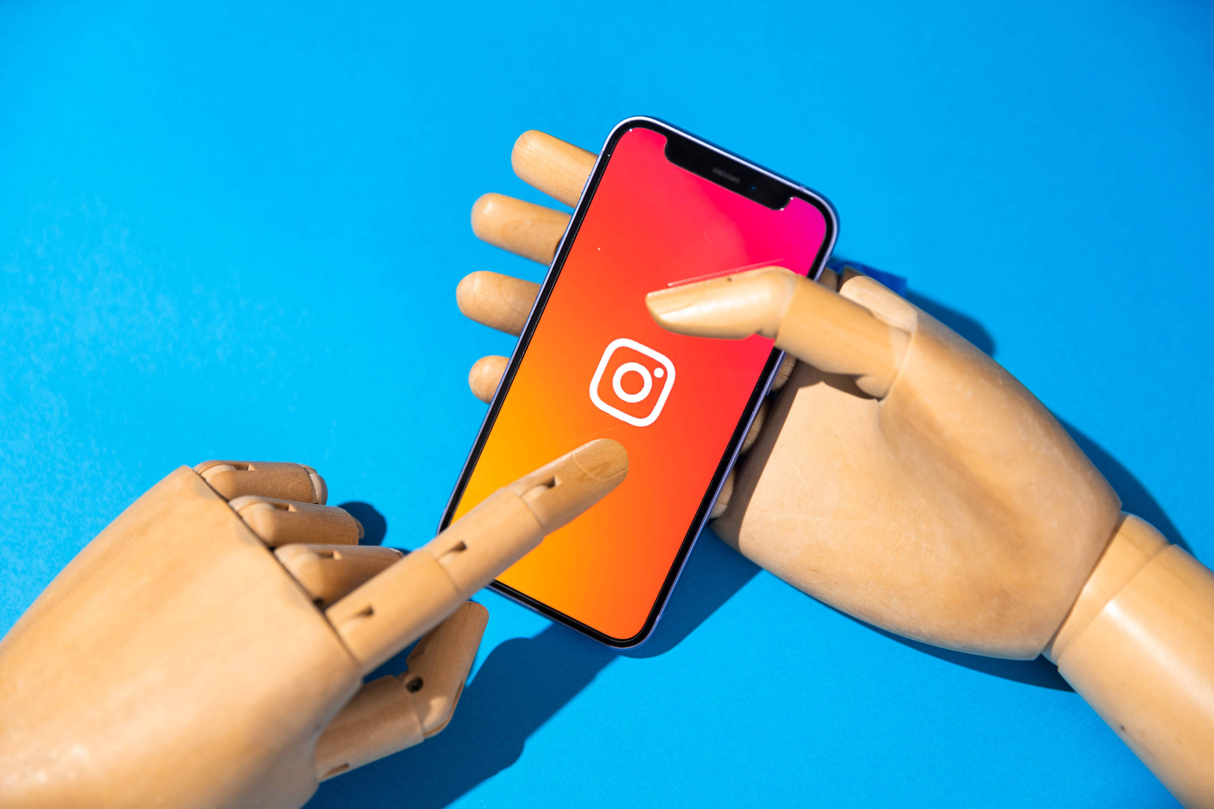 A imagem mostra duas mãos de madeira manipulando um smartphone. O celular exibe o logotipo do Instagram em uma tela com um fundo gradiente que vai do rosa ao laranja. O fundo da imagem é azul.