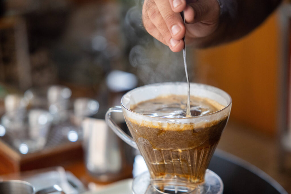 Uma mão está mexendo um coador transparente com café, que apresenta uma camada de espuma na parte superior. O fundo mostra utensílios de café e uma superfície de madeira.