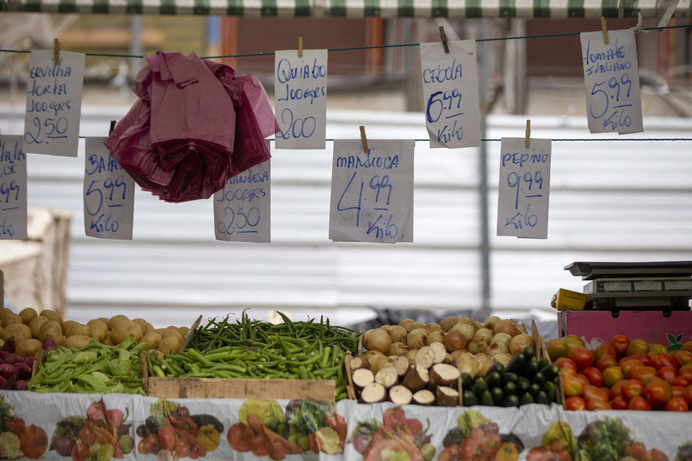 fotografia mostra barraca de feira com legumes e verduras. em um cordão de varal, acima dos alimentos, estão folha de papel sulfite indicando os preços por kilo de cada tipo