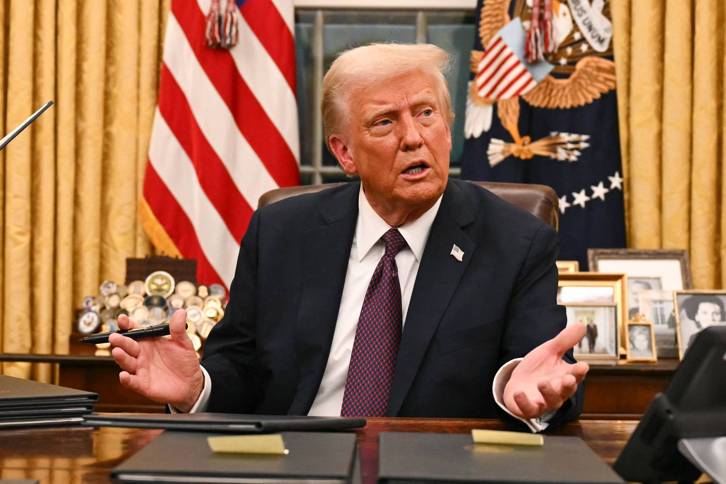 Imagem mostra Donald Trump, um homem que está sentado em uma mesa no Salão Oval, gesticulando com as mãos. Ao fundo, há cortinas douradas e bandeiras dos Estados Unidos. Em cima da mesa, há documentos e um telefone. Na parede, pode-se ver o brasão dos Estados Unidos e algumas fotos emolduradas.