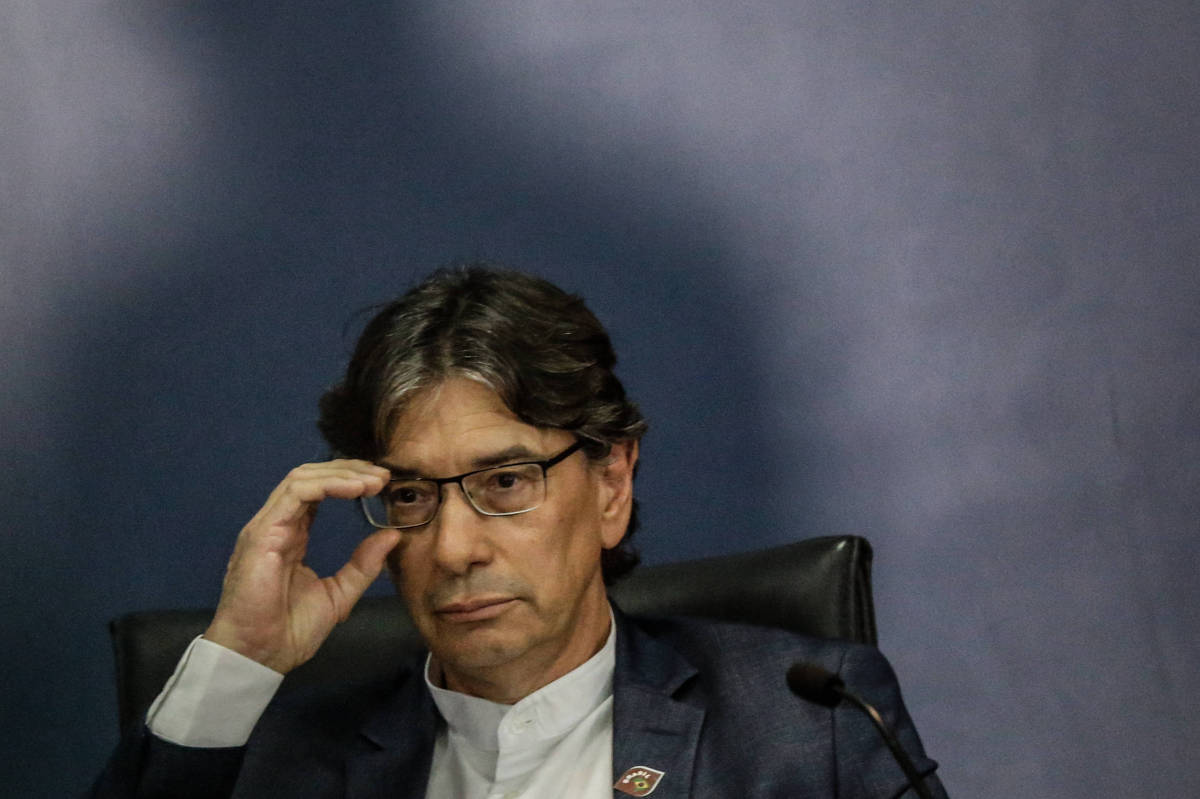 homem branco de cabelo preto, camisa branca e paletó, segura os óculos com expressão compenetrada