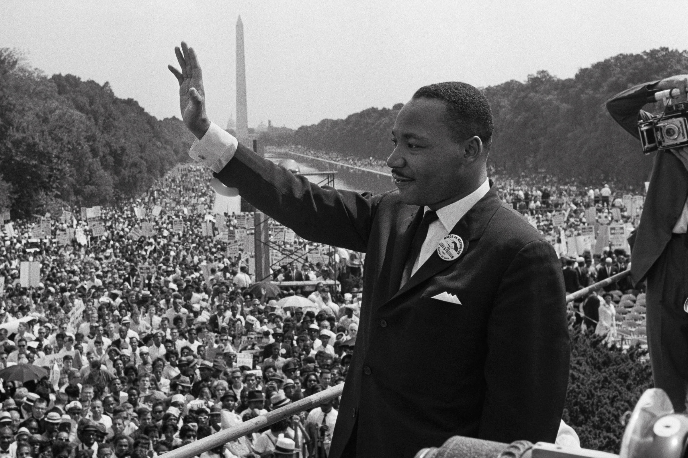 fotografia em preto e branco mostra Martin Luther King Jr em pé em um palanque. Ele é um homem negro, está vestindo terno e gravata, e acenando para uma multidão enorme em uma praça