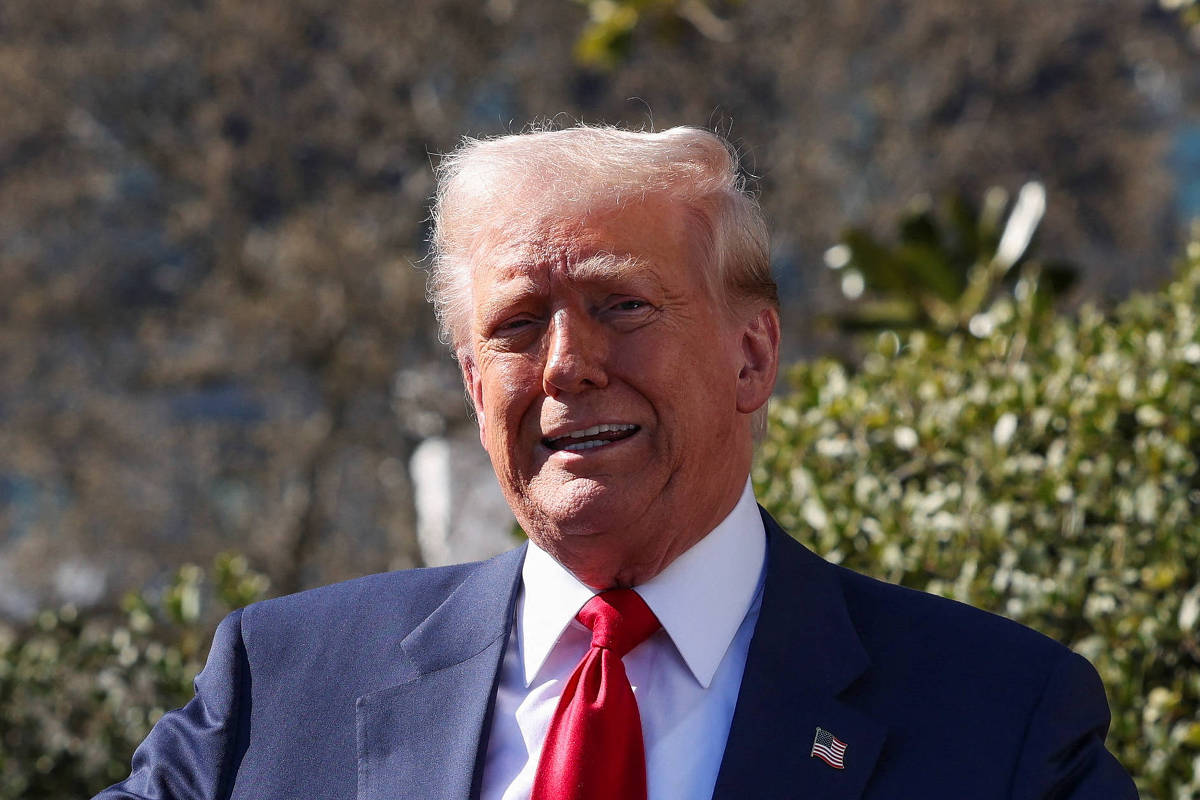 Um homem está em pé, com os braços abertos, sorrindo e segurando um papel na mão direita. Ele usa um terno escuro e uma gravata vermelha. Ao fundo, há arbustos verdes e uma estrutura arquitetônica visível.