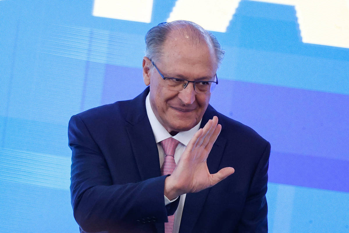 Um homem de terno escuro e gravata rosa está acenando com a mão direita. Ele usa óculos e tem cabelo grisalho. O fundo é azul com formas geométricas.