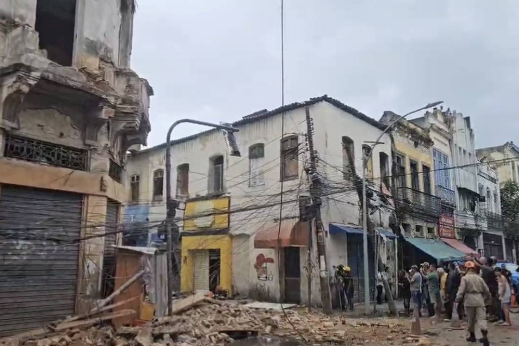 A imagem mostra uma rua em uma área urbana afetada por um desastre. Há prédios danificados, com uma estrutura parcialmente desmoronada à esquerda. Fios elétricos estão expostos e desordenados, e várias pessoas estão reunidas na calçada, algumas usando capacetes. O céu está nublado, indicando um clima sombrio.