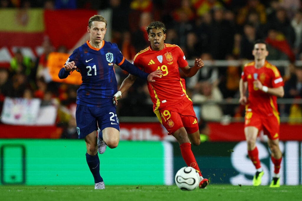 A imagem mostra um momento de uma partida de futebol entre as seleções da Holanda e da Espanha. Um jogador da Holanda, vestindo uma camisa azul escura com o número 21, está correndo em direção à bola, enquanto um jogador da Espanha, vestido com uma camisa vermelha e o número 19, está tentando alcançá-lo. Ao fundo, há torcedores e bandeiras das duas seleções.
