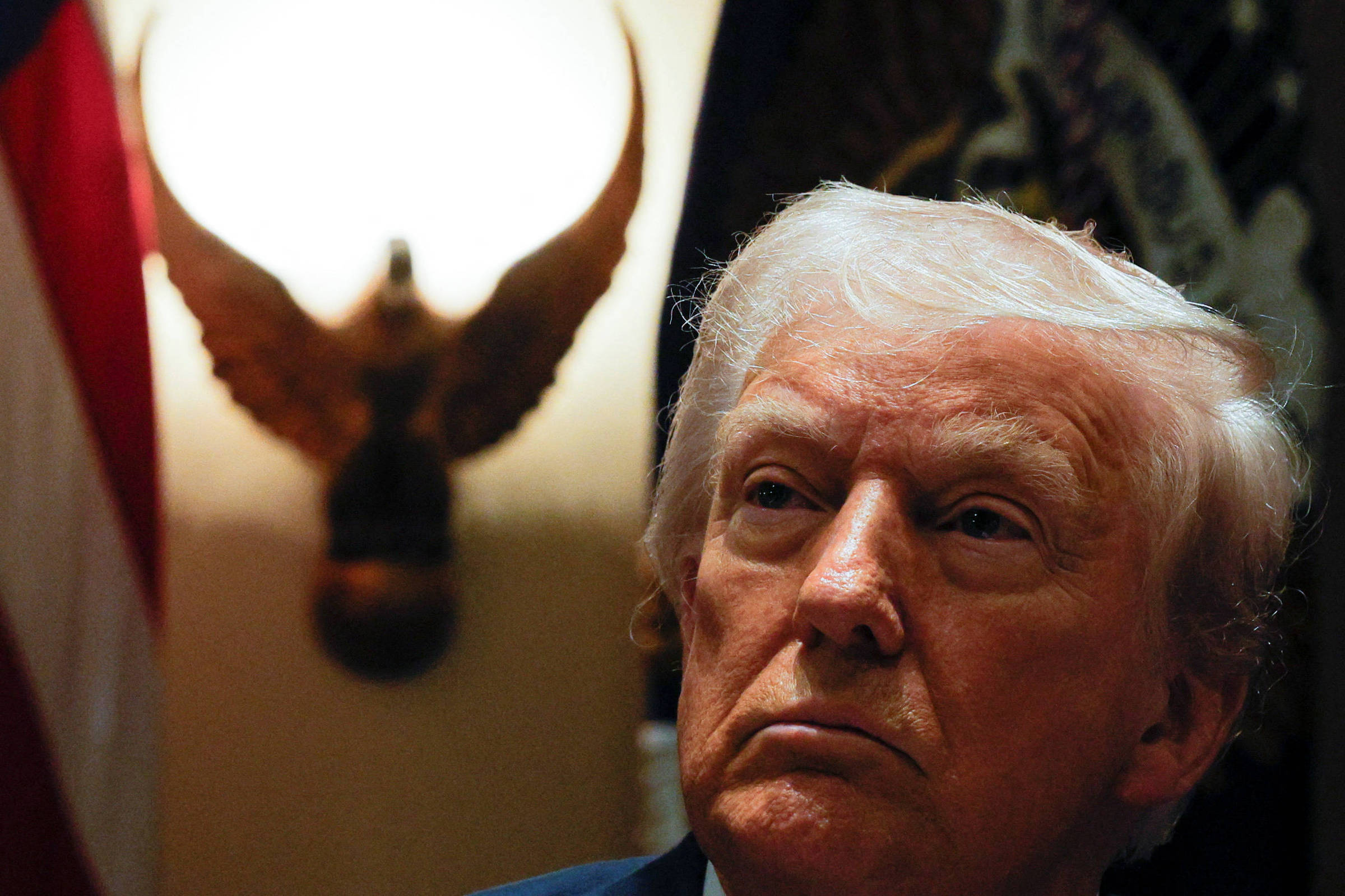 A imagem mostra um homem com cabelo loiro e uma expressão séria, em um ambiente formal. Ao fundo, há uma luz em formato circular e um símbolo de águia. A bandeira dos Estados Unidos é visível à esquerda.