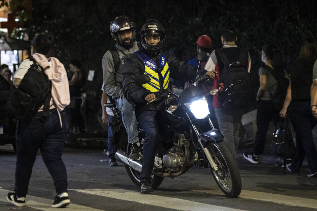 A imagem mostra uma motocicleta em movimento à noite, com dois ocupantes. O motorista está usando um capacete e uma jaqueta com detalhes em amarelo, enquanto o passageiro também está de capacete. Ao fundo, há várias pessoas caminhando na calçada, algumas delas com mochilas. A iluminação é baixa, mas a luz da motocicleta é visível.