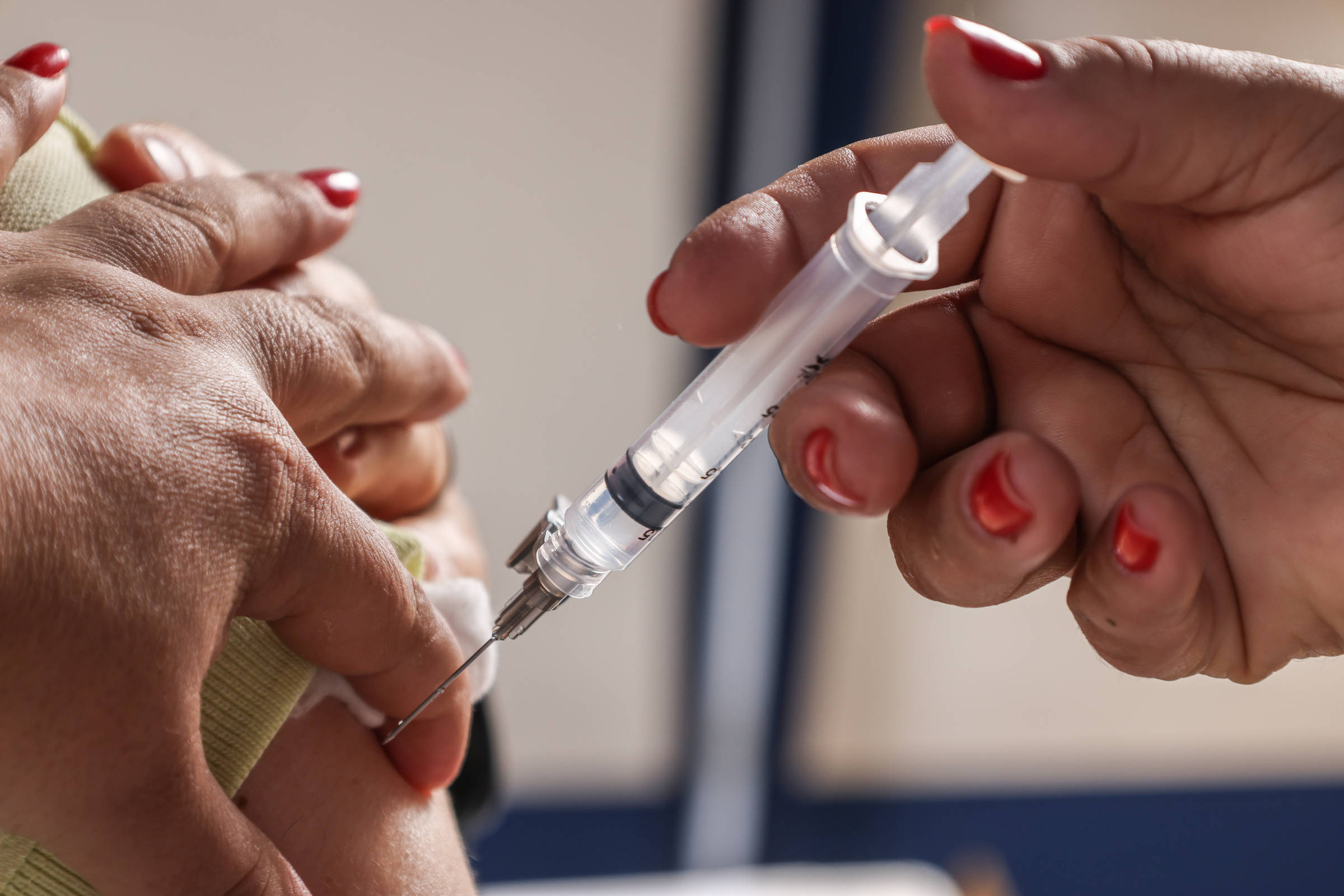 A imagem mostra uma mão segurando uma seringa enquanto outra mão está posicionada para receber a injeção. A seringa está prestes a ser inserida em um braço, que está parcialmente visível. As unhas da mão que segura a seringa estão pintadas de vermelho.