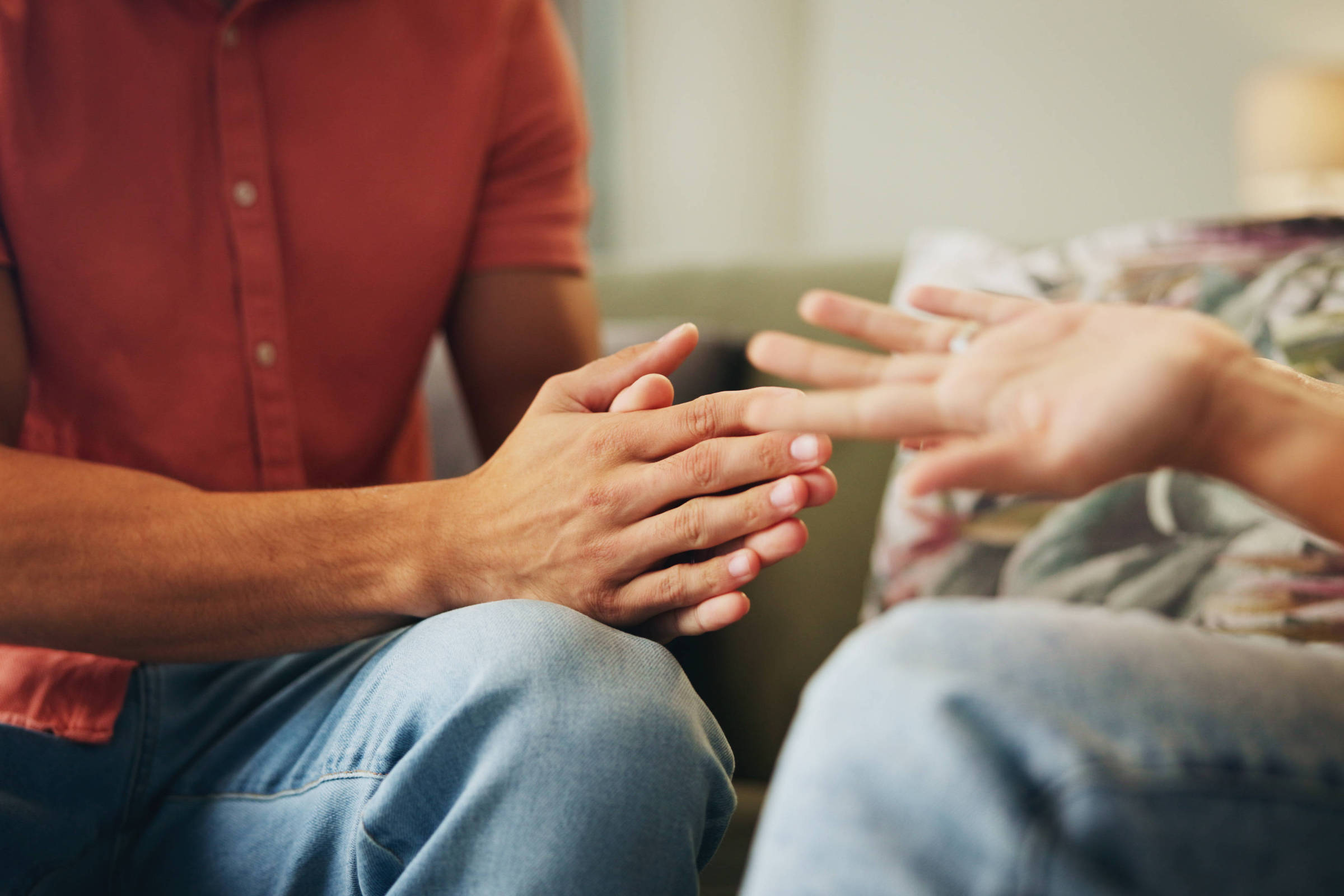 A imagem mostra as mãos de duas pessoas em uma conversa. Uma pessoa, com uma camisa de botão laranja, está com as mãos juntas, enquanto a outra pessoa, que não está totalmente visível, tem a mão estendida, como se estivesse gesticulando. O fundo é desfocado, mas parece ser uma sala de estar com um sofá e uma planta.