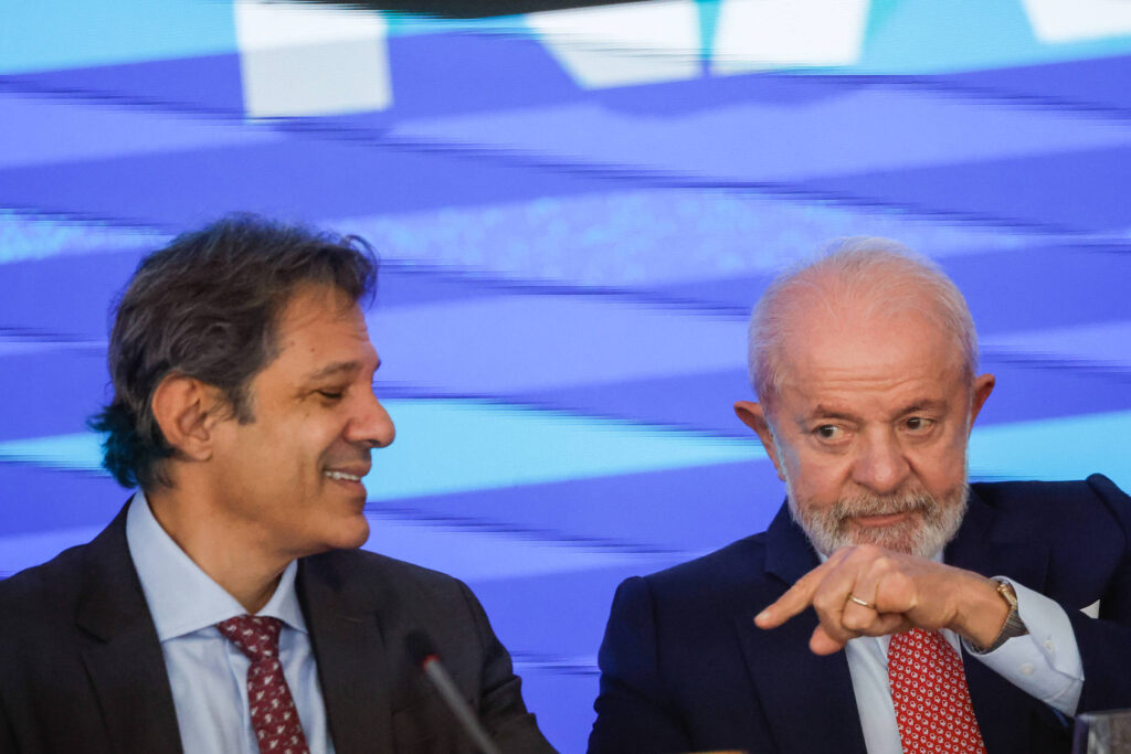 A imagem mostra dois homens sentados em uma mesa durante uma reunião. O homem à esquerda tem cabelo escuro e usa um terno escuro com uma gravata. O homem à direita tem cabelo grisalho e uma barba curta, vestindo um terno escuro e uma gravata vermelha. Eles parecem estar conversando, com o homem à direita apontando para algo com a mão.