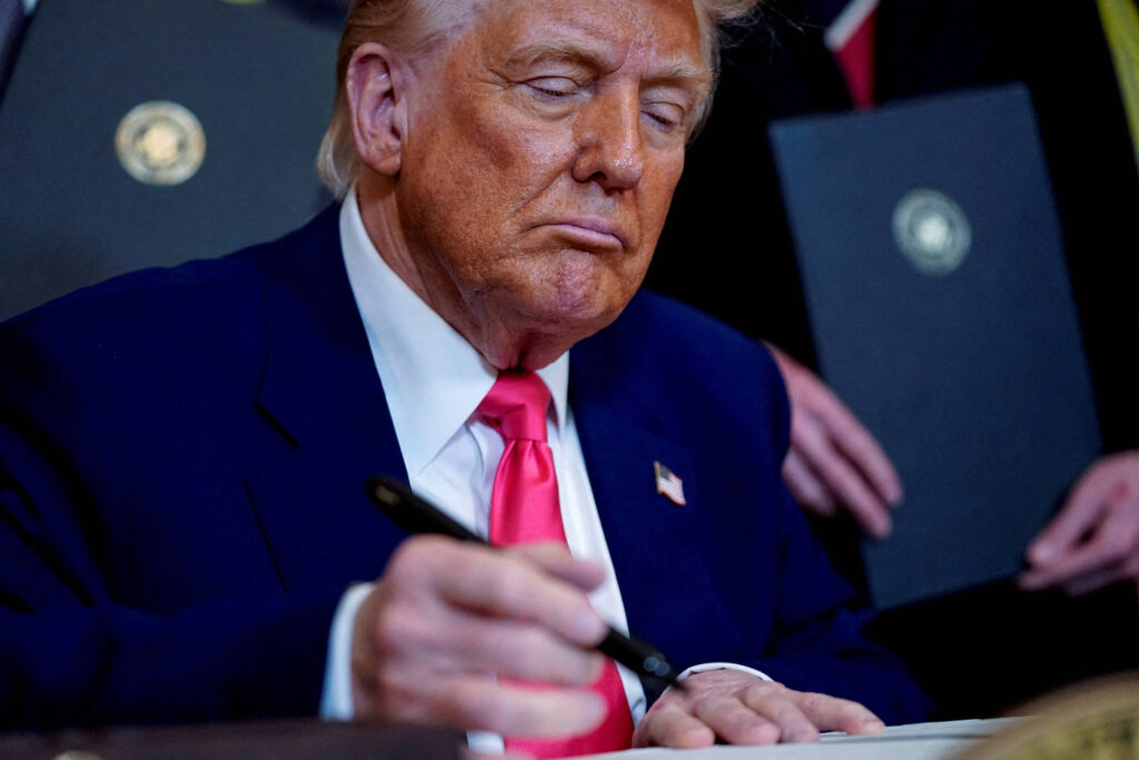Um homem com cabelo loiro e pele bronzeada está sentado à mesa, concentrado em assinar documentos. Ele usa um terno escuro e uma gravata vermelha. Ao fundo, há pessoas e pastas com o selo presidencial.